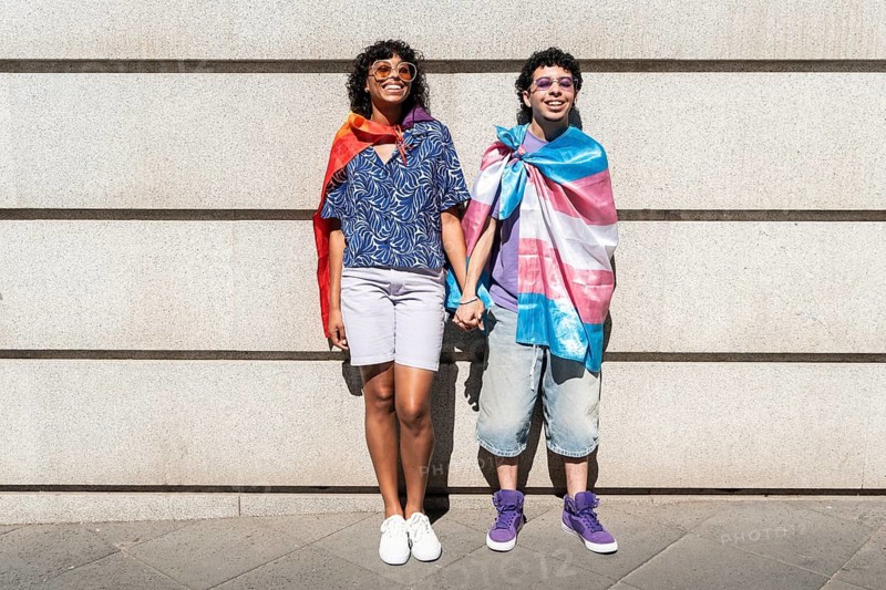 A joyful multiethnic LGBTQ+ couple stands against a wall, holding hands and wearing pride flags. They radiate happiness and unity, embodying love and diversity