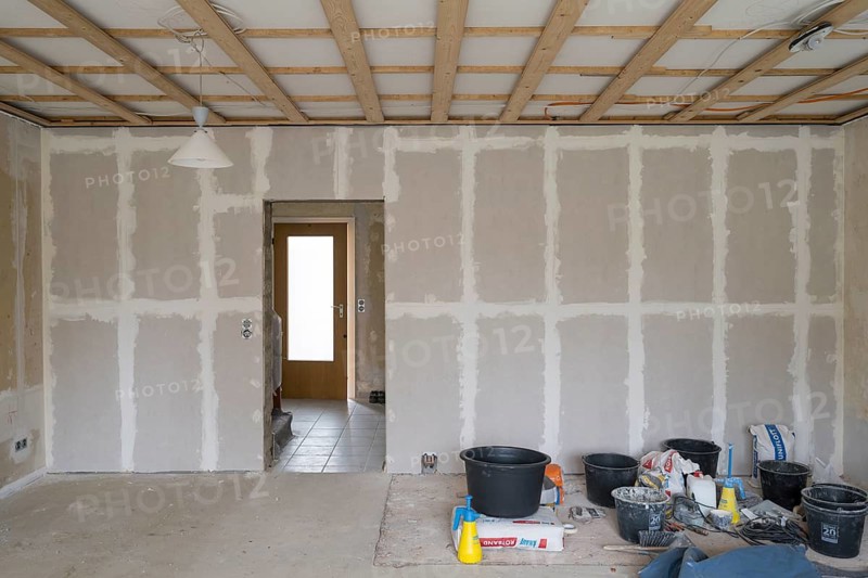 Room is being renovated, plastered wall with sheetrock, wooden structure under ceiling, preparation for ceiling panels, working materials on the floor