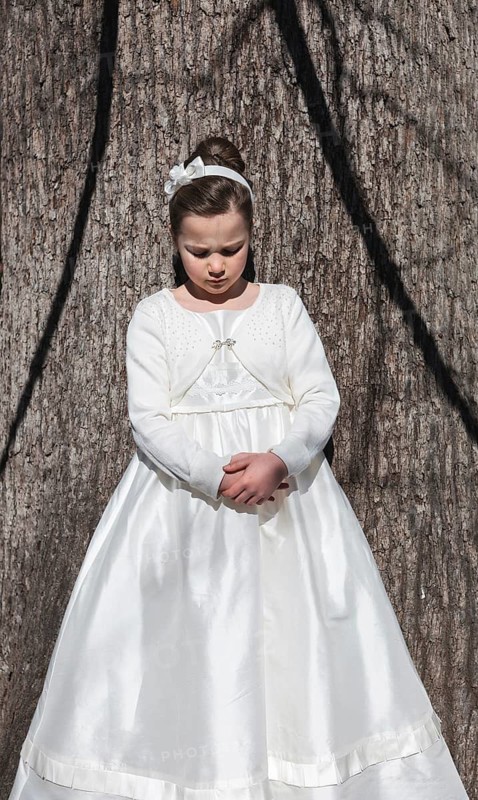 A young girl stands against a tree wearing a beautiful white First Communion dress, complete with a matching headband and a gentle expression, symbolizing innocence and celebration