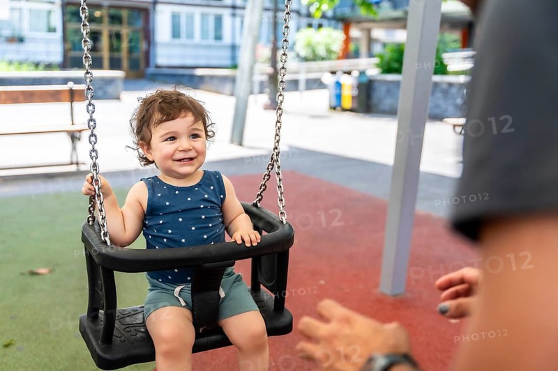 Young baby boy with curly hair smiling and laughing, sitting in a plastic swing, playing outdoors in a public park, with an adult's hands visible pushing him