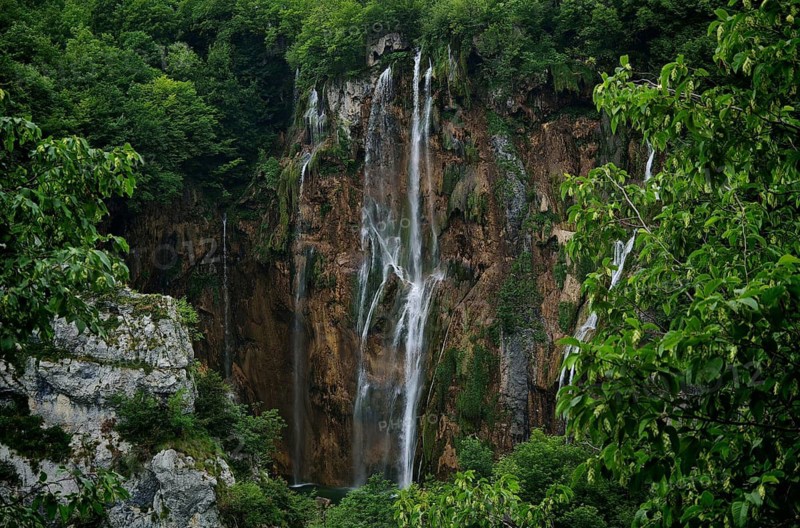 A mighty, narrow waterfall falls along a rock wall surrounded by green forest, Plitvice Lakes National Park, Croatia