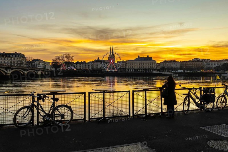 Woman stands by the river at sunset in bayonne, admiring the illuminated ferris wheel and historic skyline reflected in water, bicycles nearby during a festive christmas evening