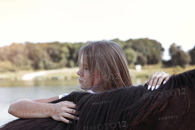 A young girl enjoys a peaceful moment with a horse by the lakeside. The serene setting features a lush green forest and calm water, perfect for relaxation and reflection