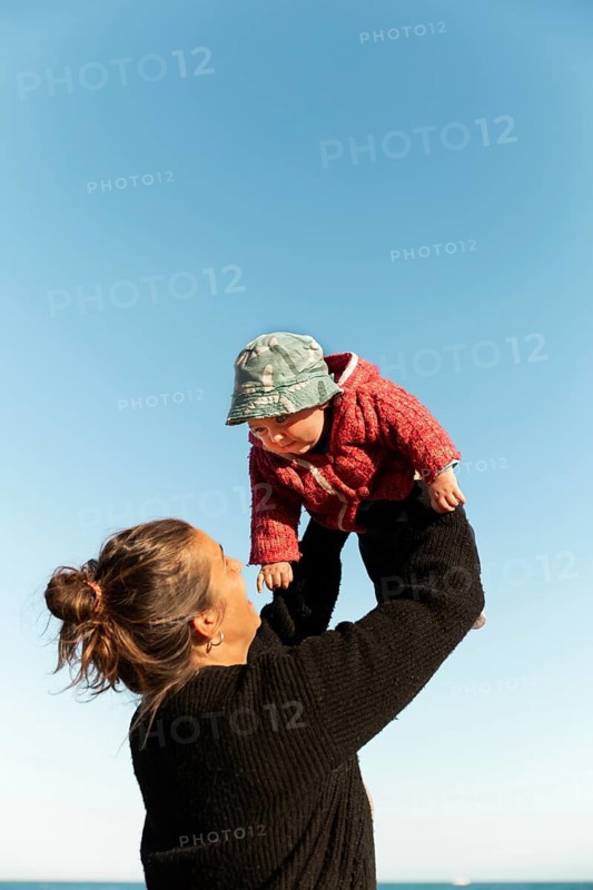 A joyful moment of a mother lifting her baby against a clear blue sky in Samalus, Barcelona. The baby wears a jacket and hat, symbolizing warmth and happiness