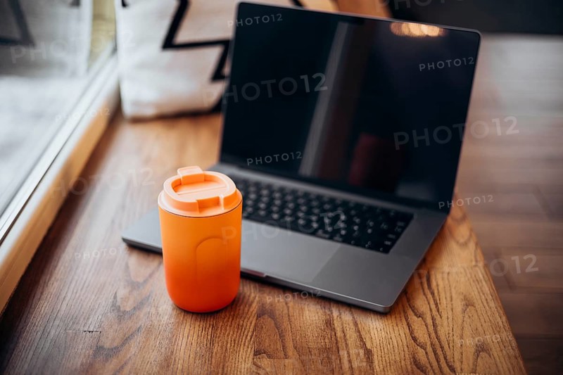 A laptop opened and placed next to an orange thermos cup on a wooden table, indicative of remote work
