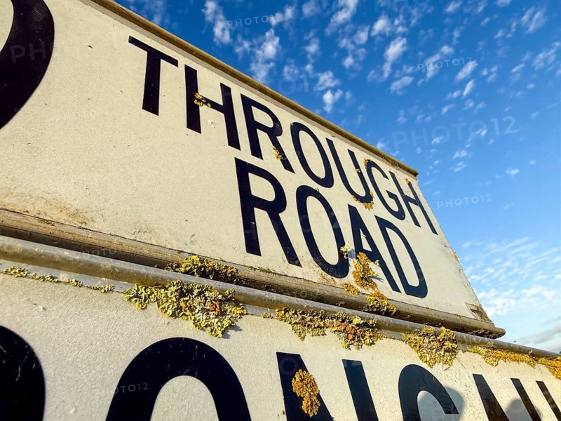 A close up of a weathered road sign with yellow lichen growing on it, set against a backdrop of a clear blue sky partially filled with white clouds in the Great Ocean Road, Australia