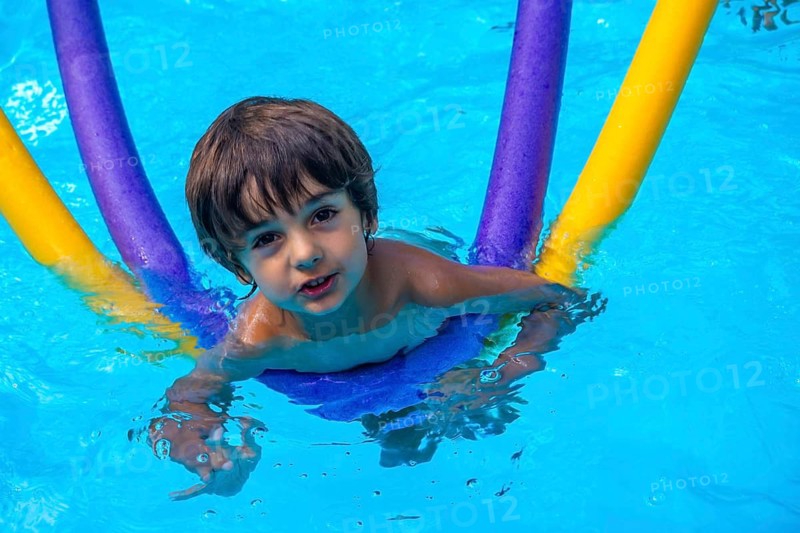 Young boy enjoying summer vacation, playing happily in a bright blue swimming pool while holding vibrant swim noodles, symbolizing childhood fun and relaxation