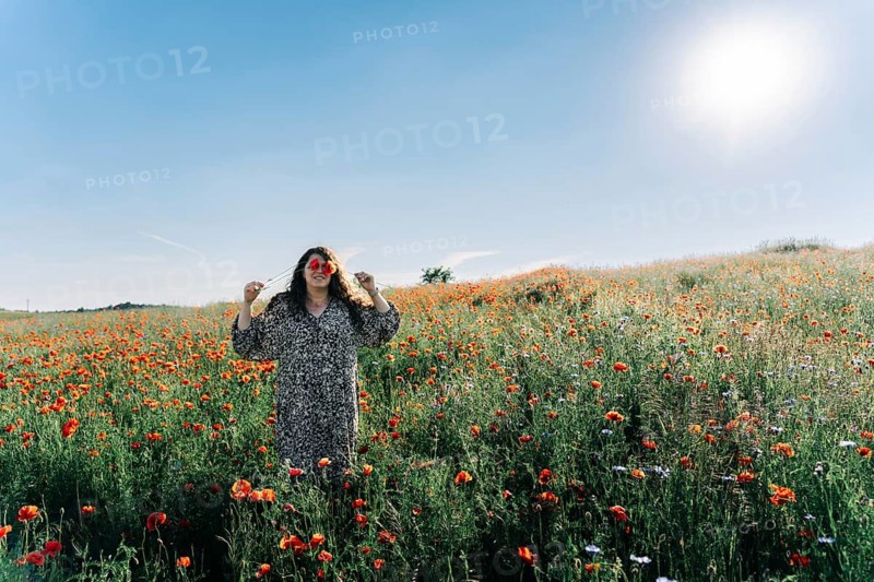 Plus size woman covering eyes with poppy (Papaver rhoeas) flowers while standing in blooming field under bright summer sun