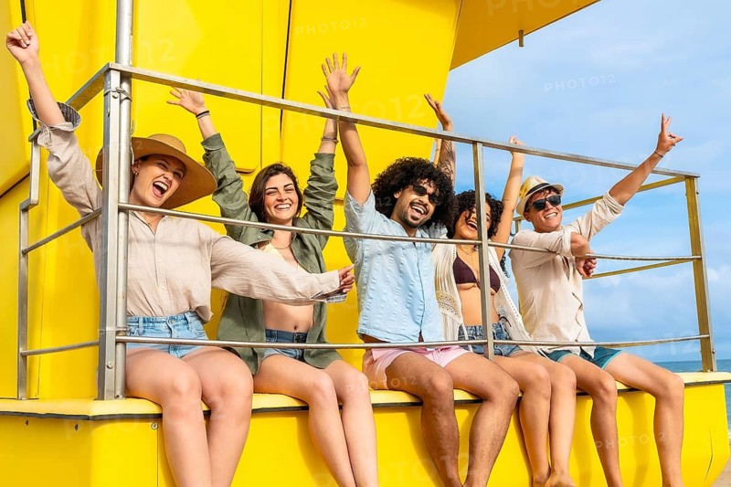 Group of happy diverse young friends sitting on a vibrant yellow lifeguard tower, cheering with raised arms while enjoying a sunny day outdoors during their summer vacation
