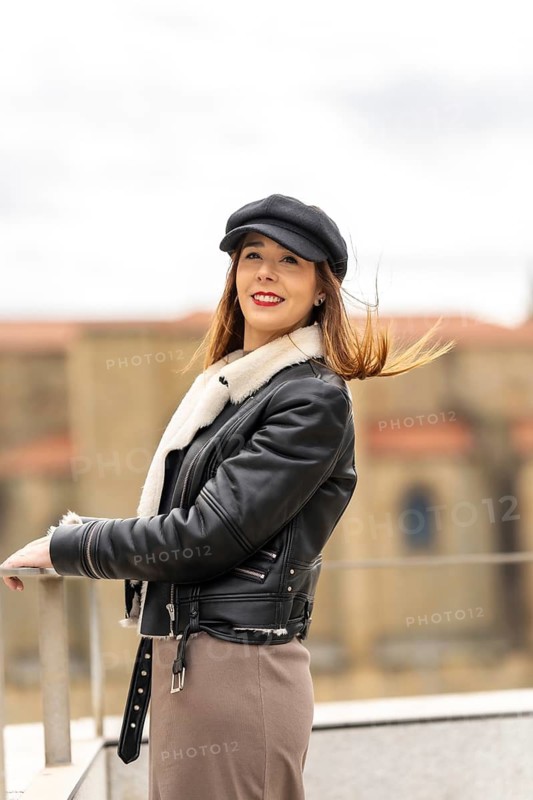 Young woman with red lipstick, wearing black cap and leather jacket, posing on a terrace with hair blowing in the wind
