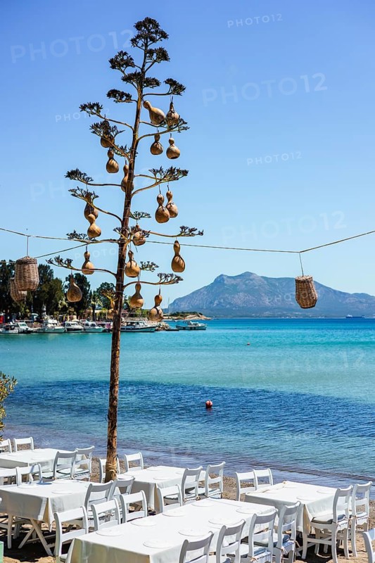 Tables lined up on the beach with turquoise water and boats in the background on a sunny day