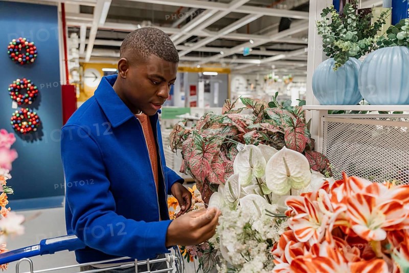 A young Black man examines artificial flowers in a shopping cart at a home decor store. He wears a blue coat and an orange tie, focused on his selection