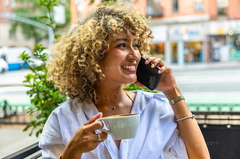 Smiling woman with curly hair talking on a mobile phone and drinking coffee at an outdoor cafe, embodying a vibrant urban lifestyle in manhattan, new york