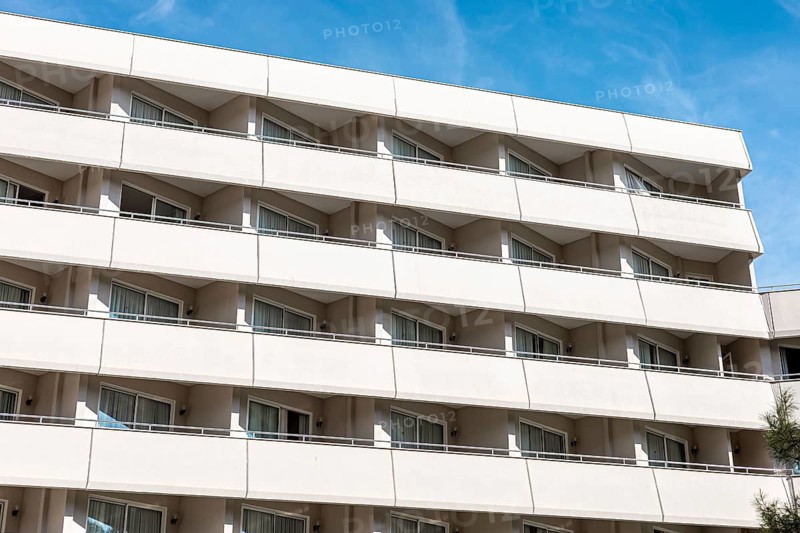 A modern apartment building with multiple balconies and large windows. The facade is light-colored, set against a clear blue sky