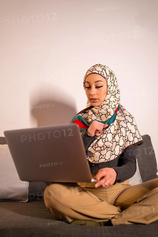 Arab girl with a white veil on her home computer on the sofa in her living room, on a video call. Young modern arab with new technologies, vertical photo