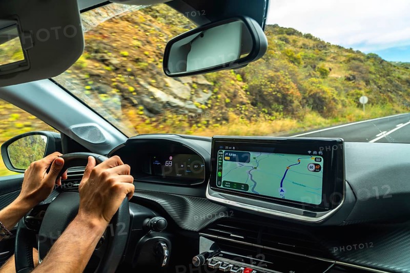 Person's hands on steering wheel while driving a modern car, with a gps navigation system on the dashboard showing a route through a mountainous, green landscape in el hierro