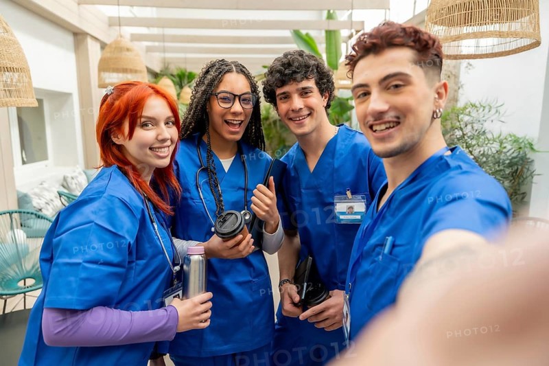 Four happy young medical students and nurse practitioners wearing blue scrubs and stethoscopes smiling at camera, capturing a cheerful group selfie moment together in a modern healthcare environment