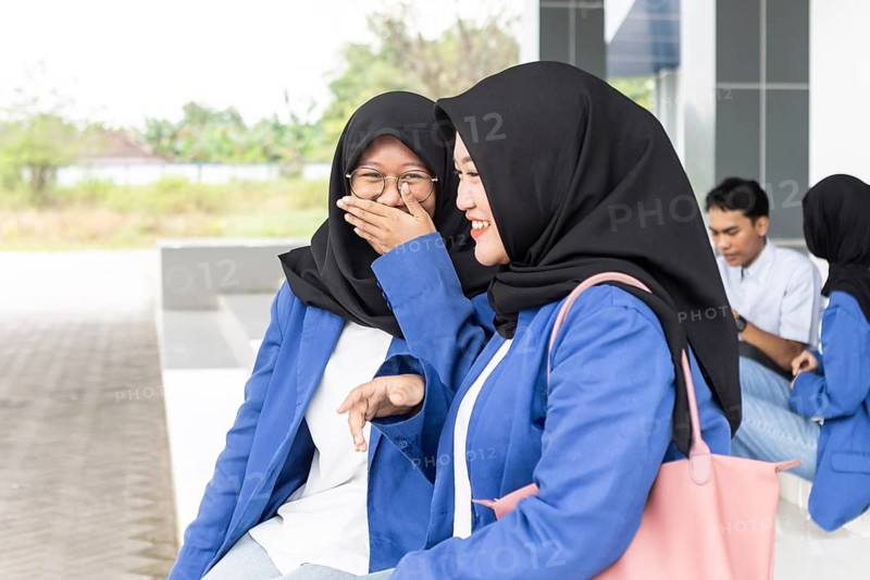 Two smiling Asian Muslim women in hijabs and blue jackets share a joyful moment while sitting outside a modern school building, symbolizing friendship and academic life