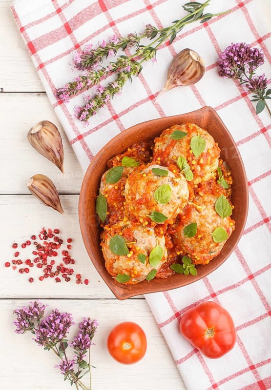 Pork meatballs with tomato sauce, oregano leaves, spices and herbs in clay bowl on a white wooden background with linen textile. top view, flat lay, close up