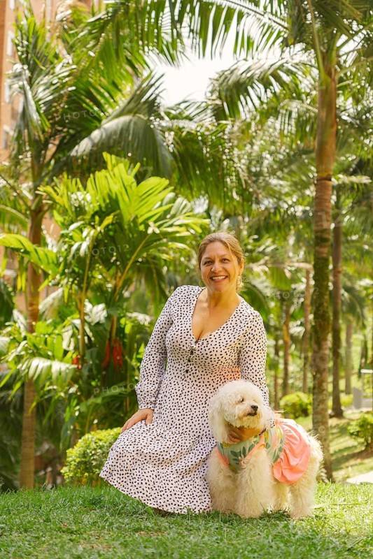 Middle-aged woman wearing a dress, kneeling on grass in a park with lush tropical vegetation, accompanied by her small white dog wearing a pink dress, enjoying a sunny day outdoors