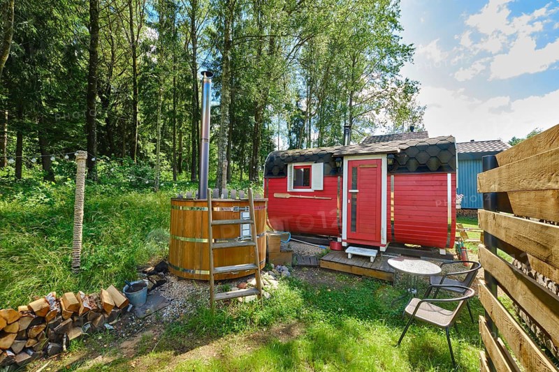 Small red sauna and hot tub in green forest surrounded by trees. Stack of firewood and chairs for visitors nearby