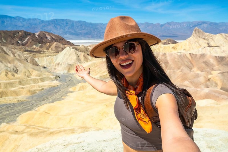 Woman in sunglasses and hat smiling and pointing toward death valley's zabriskie point badlands and distant salt flats under a clear blue sky, enjoying desert views and travel adventure