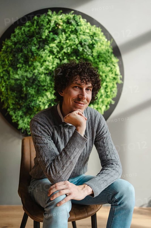 A young man with curly hair sits thoughtfully, framed by a lush green wall. This image emphasizes themes of ecology and sustainable development in a modern setting