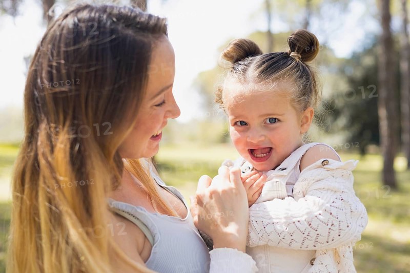 A pregnant woman lovingly embraces her young daughter, sharing a joyful moment outdoors amidst a lush, green landscape, bathed in soft, natural sunlight