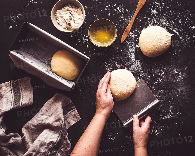 Hands gently shape dough on a floured surface, preparing it for baking. Fresh dough, flour, and a baking tin sit nearby, showing the process of creating homemade sliced bread