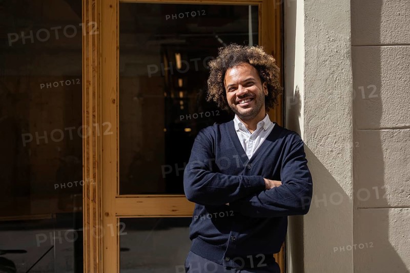A cheerful man with curly hair smiles confidently while standing outside a modern coworking space in the heart of Barcelona, showcasing the vibrant work culture of the city
