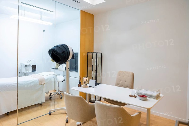 Clean and bright interior of a contemporary aesthetic clinic, featuring a private consultation desk in the foreground and a treatment bed with advanced equipment visible through a glass partition