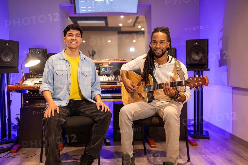 Two musicians in a modern recording studio. One playing an acoustic guitar, the other seated. The environment is filled with high-tech audio equipment and soft lighting