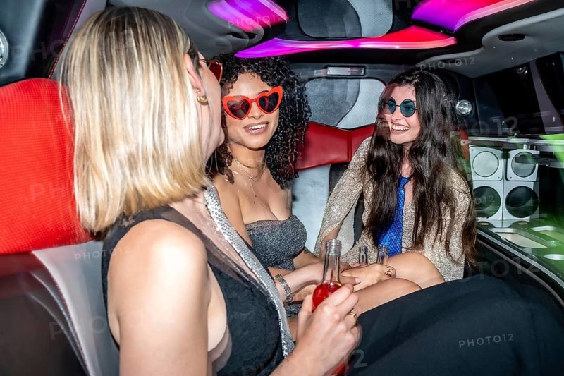 Three diverse young women enjoying a night out, laughing and holding drinks inside a luxurious limousine with vibrant neon lights, embracing celebration and friendship