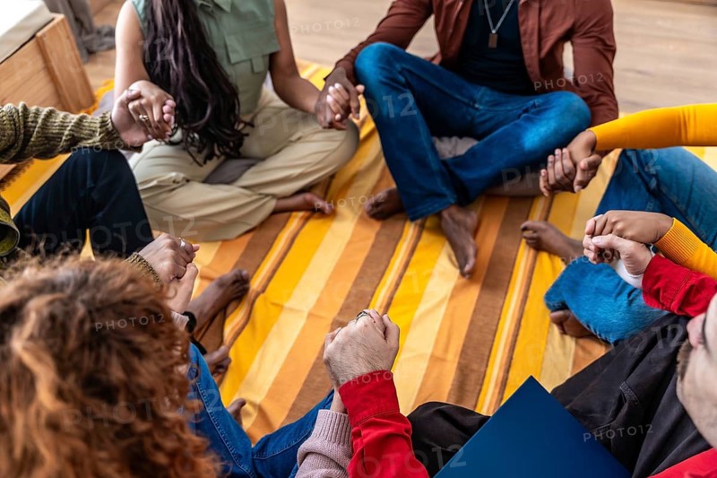 Diverse group sitting on a blanket in a circle, holding hands and sharing support during a therapy or support meeting, expressing connection, empathy, healing and trust