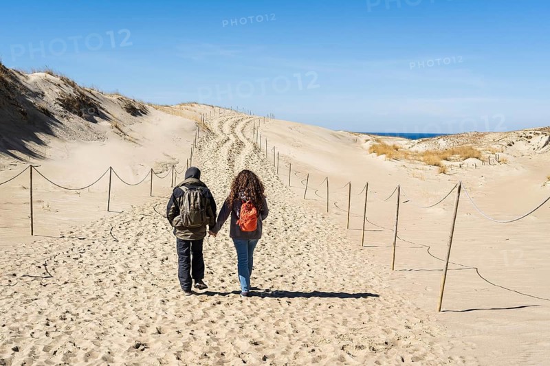 A couple of backpackers walk along a hiking trail in sand dunes on the Curonian Spit near the Baltic Sea, A couple walking hand in hand along a sandy path through dunes under a clear blue sky, Curonian Spit, Neringa, Lithuania