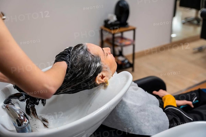 Stylist in black gloves shampooing and massaging a client's dark hair at a salon basin, creating foamy lather for a relaxing, professional hair care and pampering treatment