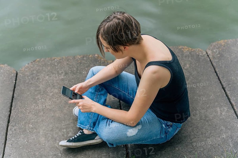 Overhead view of woman 40s using smartphone on urban riverbank, A woman in casual attire sits on concrete by the water, engrossed in her smartphone
