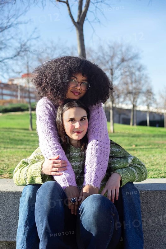 Two friends sitting outdoors on a sunny day, sharing a warm embrace. They are wearing colorful sweaters and enjoying each other company in a peaceful park setting