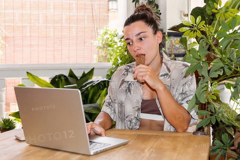 A woman enjoys remote work from a laptop in a tropical-themed environment, savoring an ice cream treat while surrounded by lush green indoor plants, during her vacation