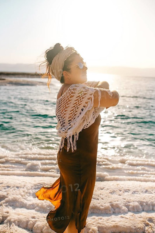 Side view of woman standing at the Dead Sea in Jordan, bathed in the sunset sunlight while wearing a stylish outfit with bohemian influences
