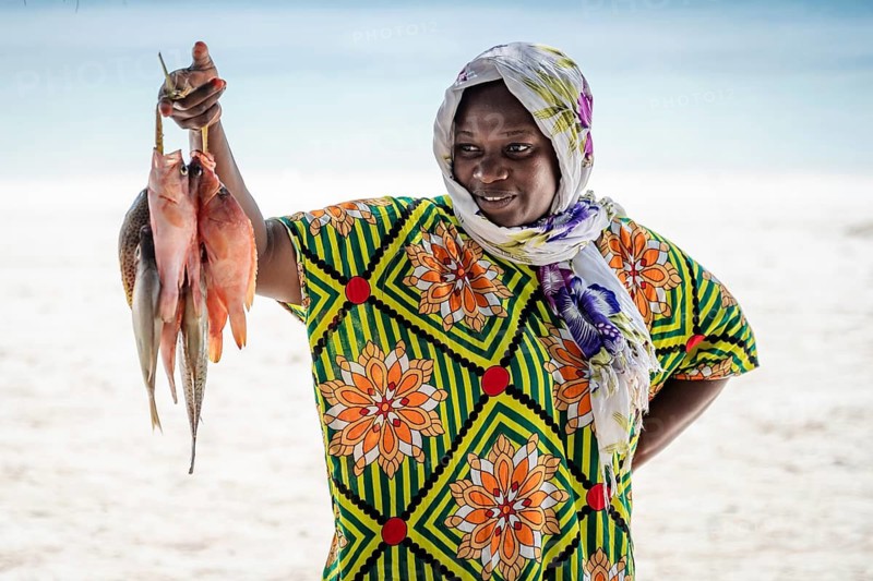 MICHAMVI, ZANZIBAR - OCTOBER 15, 2025: A local African woman in a colorful traditional dress holding up a bunch of freshly caught fish on a white sand beach