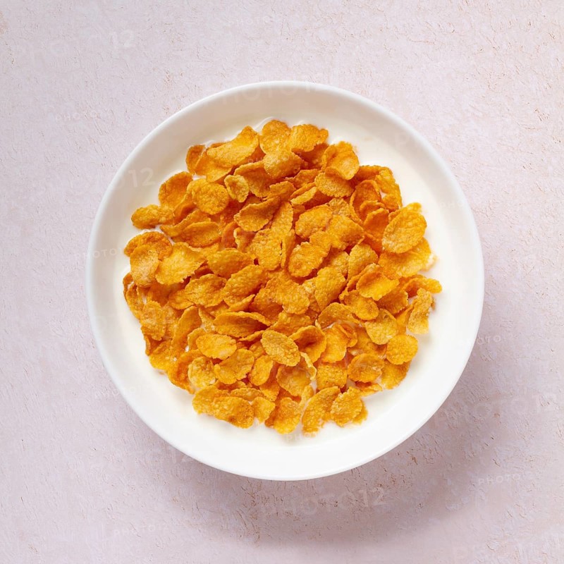 Top view of a white bowl full of crunchy golden cornflakes, isolated on a light textured background. Perfect for a healthy and quick breakfast option