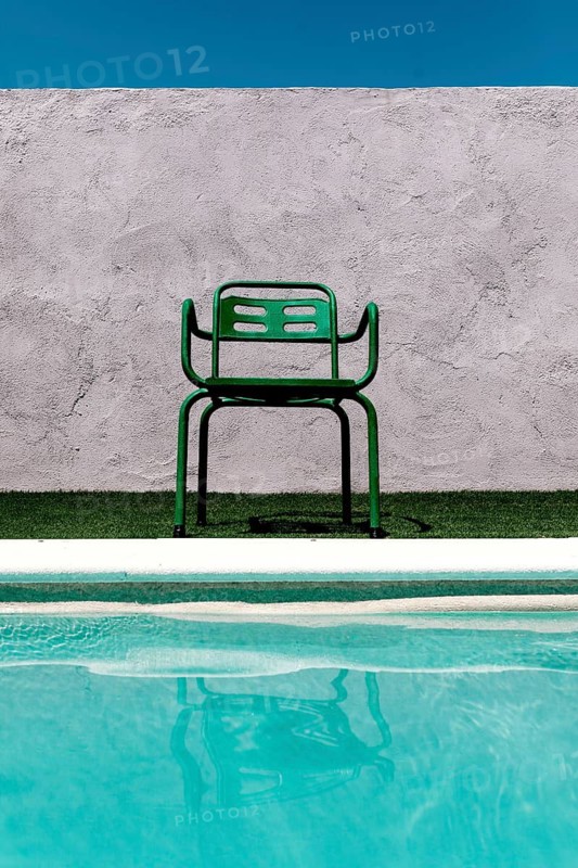 A vibrant green chair sits on grass by a pool, reflected in the water. Above, the clear blue sky complements the textured, light gray wall backdrop, creating a serene summer scene