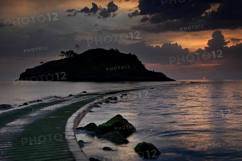 Silhouette of a small rocky island in the Atlantic Ocean, onto which a half-flooded causeway leads, at sunrise and dramatic clouds, Basque Country, Spain