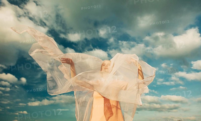 A woman is wrapped in sheer, flowing fabric against a vast, cloudy sky. The fabric catches the light, creating a dreamlike and ethereal atmosphere