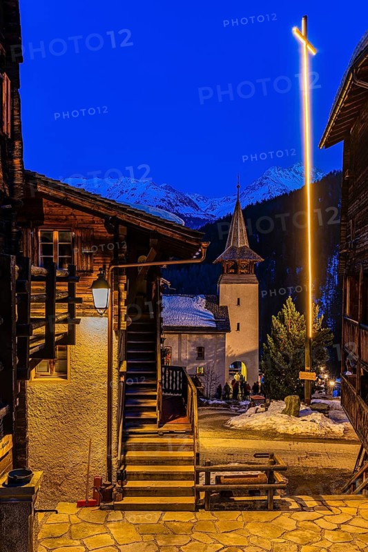 Illuminated Saint Theodule church and illuminated large wooden cross, night view, historic town center, Grimentz, Val d'Anniviers, Valais Alps, Canton of Valais, Switzerland