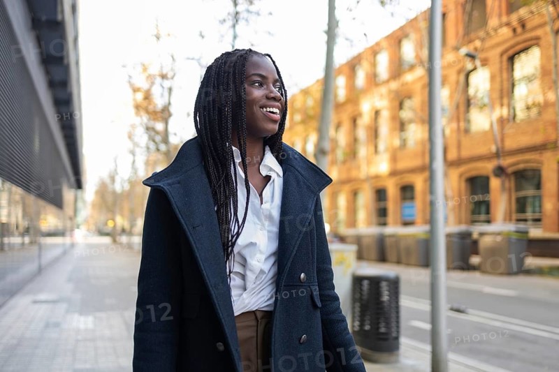 An African American businesswoman with braids, wearing a formal coat over business attire, enjoys walking in an urban setting