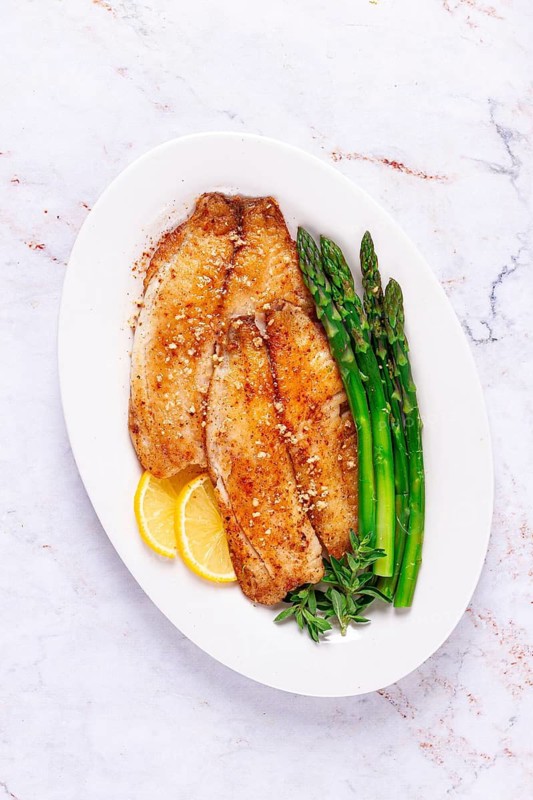 Fresh fish filets are served on a white plate alongside green asparagus and slices of lemon. The dish is prepared in a kitchen environment, ready to eat