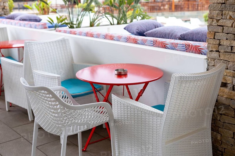 Brightly colored outdoor seating features white chairs and a red table, creating a cheerful atmosphere. Cushions and plants enhance the inviting space for relaxation. Summer terrace cafe