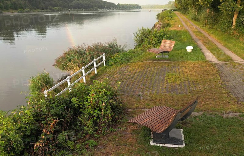 A quiet river with green riparian vegetation and several wooden benches to relax on, ergonomic wooden loungers on the Kiel Canal, NOK, Kiel Canal, Schleswig-Holstein, Germany